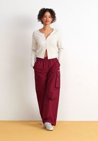 White ribbed cardigan with a button front, paired with loose burgundy cargo pants featuring side pockets and a small logo on the leg.
