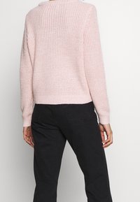 Hellrosa gestrickter Pullover mit strukturierter Oberfläche, cropped Design und langen Ärmeln. Kombiniert mit schwarzen, eng anliegenden Hosen.