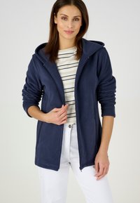 Chaqueta de forro polar azul marino con cierre de cremallera, capucha y puños acanalados, llevada sobre una camiseta de rayas y combinada con pantalones blancos.