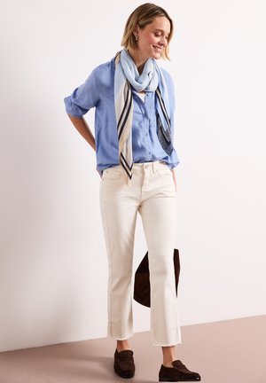 Frau trägt hellblaues Hemd mit Knöpfen, gemusterten Schal, weiße gekürzte Hose und dunkelbraune Loafer, steht vor einfachem Hintergrund.