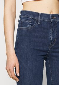 Jeans de denim azul oscuro con una forma ajustada, que presentan un diseño de cinco bolsillos, costuras visibles y un cierre de botón en la cintura.