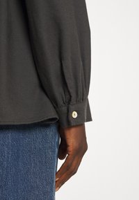 Sort langærmet skjorte med samlet stof ved håndleddet og en knapdetalje. Kombineret med blå denim jeans. Blød tekstur generelt.