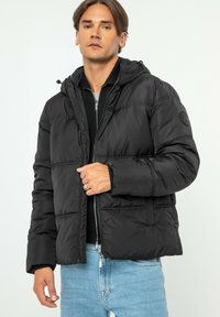 Černá péřová bunda s kapucí, zapínáním na zip a elastickými manžetami; má šité panely a kulaté logo na levém rukávu.