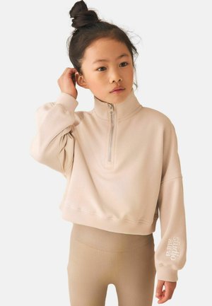 Young girl with dark hair in a top bun wearing a beige zip-up sweatshirt and ribbed beige leggings, touching her ear thoughtfully.