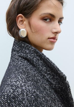 Veste à motif en chevron noir et blanc avec un col haut. Le mannequin porte des boucles d'oreilles rondes en argent et a les cheveux bruns en désordre.