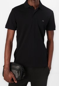 Polo noir en coton avec col et manches courtes, arborant un petit logo sur la poitrine. Associé à un sac noir texturé.