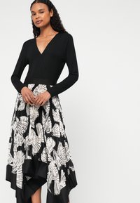 Μαύρο φόρεμα με μακριά μανίκια και βαθύ V-λαιμό, με φούστα σε πτυχωτό μαύρο και λευκό σχέδιο φύλλων και εφαρμοστή μέση.