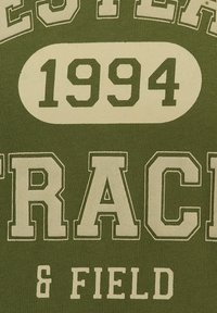 Tessuto verde oliva con grande testo stampato in bianco che legge "WESTERN TRACK & FIELD" e "1994" in un carattere audace e atletico.
