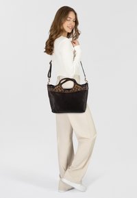 Donna con capelli lunghi e mossi, indossa una maglietta bianca e pantaloni beige, mentre porta una borsa a tracolla in pelle scura con borchie decorative dorate, sorridendo con gli occhi chiusi.