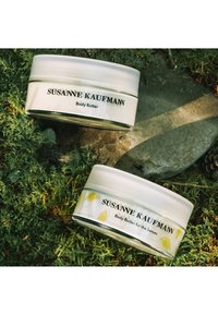 Deux bocaux ronds blancs de beurre corporel avec des couvercles transparents, étiquetés "SUSANNE KAUFMANN." L'un présente des illustrations d'oiseaux jaunes, reposant sur de la mousse.