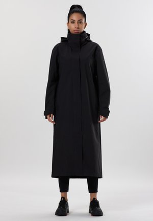 Cappotto da pioggia lungo e nero con colletto alto, realizzato in materiale impermeabile. Presenta una linea dritta ed è indossato sopra leggings neri e sneakers.