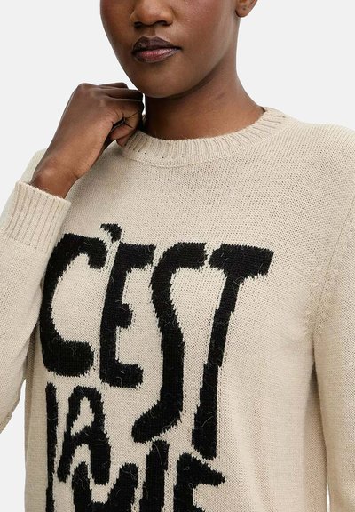 Maglione a maglia beige con scollo rotondo caratterizzato da un prominente design grafico di testo nero. Texture morbida e polsini a costine visibili.