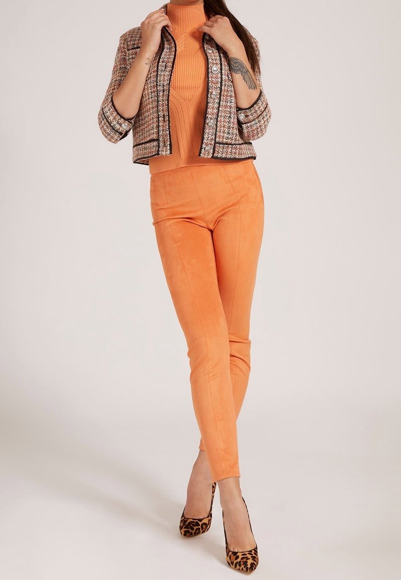 Femme portant un col roulé orange et un pantalon avec une veste en tweed et des talons hauts imprimé léopard, debout devant un fond uni.