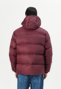 Chaqueta de plumas burdeos con capucha, diseño cuadrado, textura acolchada y puños elásticos. Combinada con vaqueros de mezclilla azul claro.