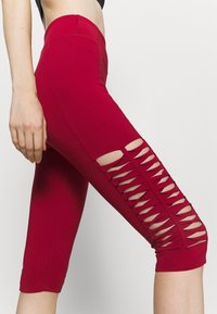 Rote Capri-Leggings aus elastischem Stoff, mit einem Ausschnittdesign an den Seiten und eng anliegenden Bündchen am Saum. Glatte Textur.