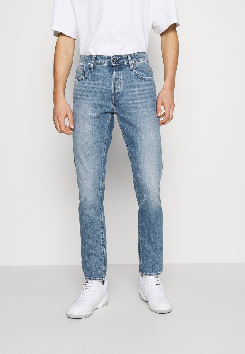 Homme portant un jean slim bleu délavé avec de légères marques d'usure, un t-shirt blanc et des baskets blanches, debout devant un fond uni.