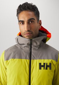 Chaqueta amarilla brillante y gris con un alto cuello, que presenta un logo negro y acentos naranjas en la capucha. Textura suave, cierre de cremallera.