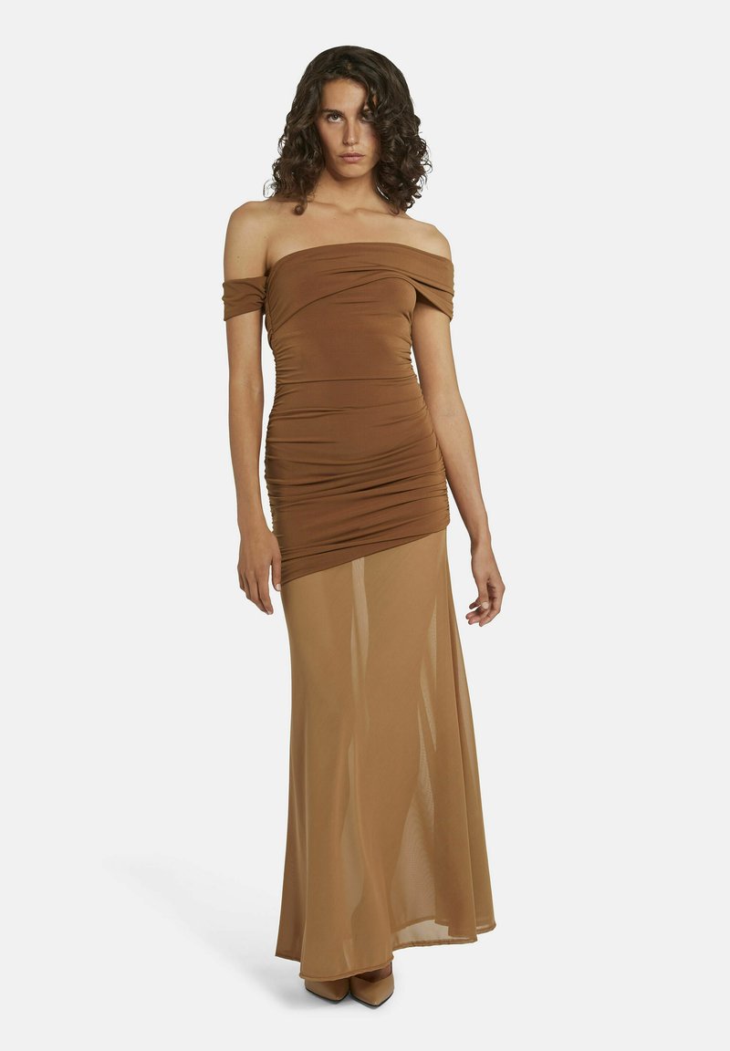 Vrouw met krullend haar in een bruine jurk met een off-shoulder en een strakke pasvorm, met een doorschijnende, lange rok en puntige hakken, staande tegen een witte achtergrond.