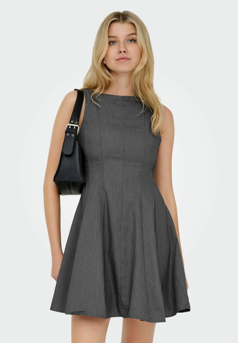 Donna bionda che indossa un vestito grigio scuro senza maniche, mentre tiene una borsa a tracolla nera su uno sfondo uniforme.