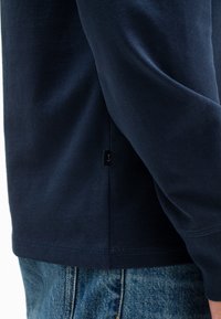 Navyblaues Langarmhemd mit glatter Textur, figurbetontem Schnitt und subtiler Etikettendetails an der Seite, kombiniert mit Jeans.