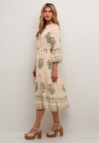 Long, beige dress with green floral patterns, tiered design, and three-quarter bell sleeves. Tied waist and lace accents at the hem.