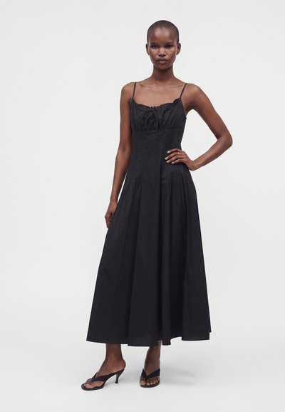 Femme portant une longue robe noire sans manches avec de fines bretelles et des sandales noires à talons, debout avec une main sur la hanche.