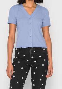 Top corto a maniche corte di colore azzurro chiaro a costine con scollatura a V, dettaglio di bottoni e orlo frill, abbinato a pantaloni neri con ricami floreali bianchi.