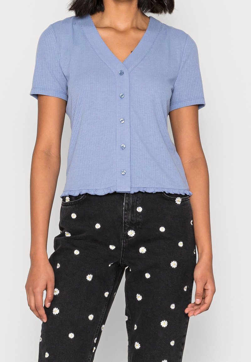 Top corto a maniche corte di colore azzurro chiaro a costine con scollatura a V, dettaglio di bottoni e orlo frill, abbinato a pantaloni neri con ricami floreali bianchi.