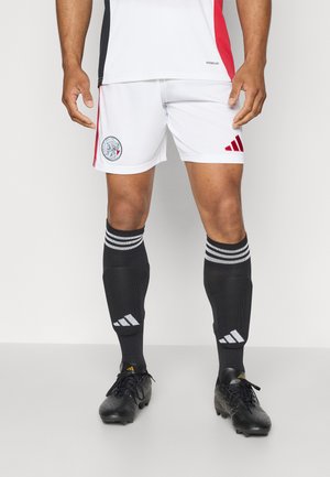 Pantaloni corti da calcio bianchi con dettagli rossi e logo, abbinati a calze nere con strisce bianche e scarpini neri. Tessuto di finitura liscia.