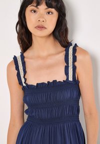 Vestido azul marino con tirantes rizados y corsé fruncido, que presenta adornos de encaje en los tirantes y una tela fluida con textura.