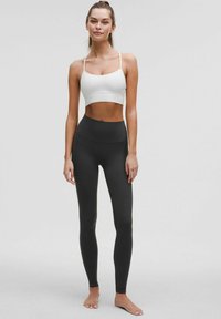 Valkoinen urheiluliivi ohutolkaimilla yhdistettynä korkeavyötäröisiin mustiin leggingseihin, molemmat sileitä ja venyviä materiaaleja neutraalia taustaa vasten.