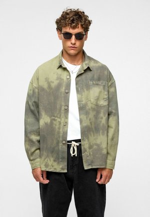 Groene tie-dye overshirt met een knopenfront, borstzak en relaxed fit, gecombineerd met een wit t-shirt en zwarte broeken.