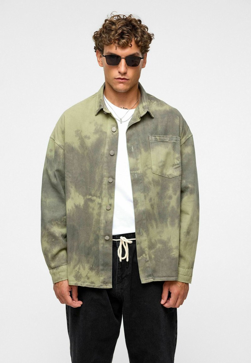 Groene tie-dye overshirt met een knopenfront, borstzak en relaxed fit, gecombineerd met een wit t-shirt en zwarte broeken.