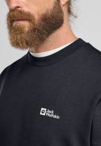 Felpa grigia scura con colletto a girocollo, caratterizzata da un logo bianco di Jack Wolfskin sul petto. Tessuto liscio con una consistenza morbida.