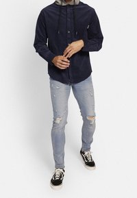 Camisa de botones de pana azul marino con capucha gris, combinada con jeans skinny desgastados de color azul claro y zapatillas negras con cordones blancos.
