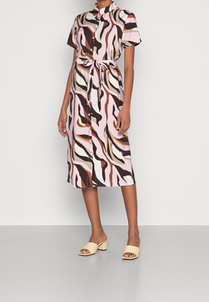 Kvinde iført en kjole med korte ærmer og knapper foran i midi-længde med pink, brun, orange og hvide abstrakte mønstre samt cremefarvede blokhælede sandaler med åbne tæer.