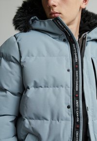 Veste matelassée bleu clair avec une capuche doublée de fourrure noire, dotée d'un zip à l'avant, de poches latérales et d'un passepoil logo contrasté le long du zip.