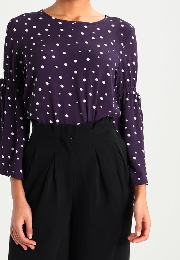 Blouse violette à pois avec manches longues à volants, associée à un pantalon noir taille haute avec plis et fermeture éclair à la taille.