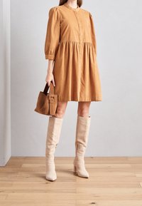 Abito in velluto a costine marrone a maniche corte, con colletto rotondo e arricciature in vita, abbinato a stivali beige alti fino al ginocchio con tacco a spillo e una piccola borsa.