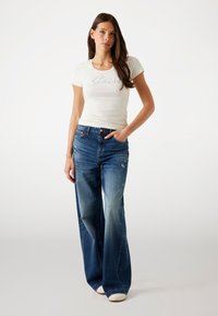 Maglietta bianca aderente con logo "Guess", abbinata a jeans a gamba larga blu con lavaggio sbiadito e lievi abrasioni, completata da sneaker bianche.