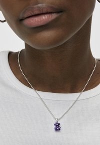 Collar de cadena de bolas de plata presenta un colgante en forma de oso morado con un acabado brillante. Una camiseta blanca de canalé simple sirve como fondo.