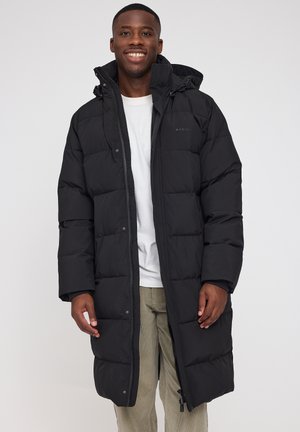 Schwarze gepolsterte Jacke mit Reißverschluss und Druckknopfverschluss, mit Kapuze und seitlichen Taschen. Getragen über einem weißen T-Shirt und hellen Hosen.