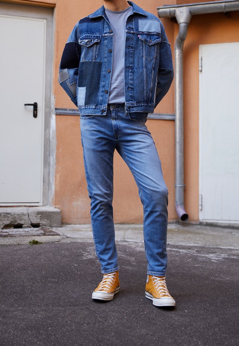 Homme portant une veste en jean patchwork, un jean bleu clair et des baskets montantes jaunes, debout à l'extérieur près des portes.