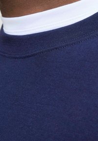 Tamno plava pamučna tkanina s glatkom teksturom, s bijelim rebrastim ovratnikom. Prikaz izbliza naglašava detalje šivanja.