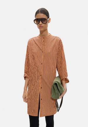 Kobieta w dużej pomarańczowo-czarnej koszuli w pionowe paski, duże okulary przeciwsłoneczne, trzyma złożoną zieloną kurtkę i czarną torbę.