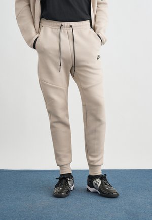 Persona che indossa pantaloni jogger beige Nike con coulisse nere, maglietta nera e scarpe sportive nere in piedi su un tappeto blu.