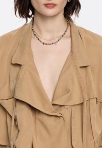 Beige oversized jacket met een gedrapeerde kraag, voorzien van een zachte textuur en een rits aan de zijkant. Gecombineerd met een multi-kleurige kralenketting.