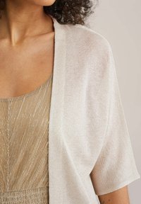 Světlé béžové šálo s krátkými rukávy, vyrobené z měkké, lesklé látky. Elegantně přiléhá přes metalické zlaté šaty se vzorem chevron