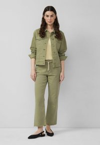 Mujer con cabello largo, oscuro y rizado, vistiendo una chaqueta verde oliva, pantalones anchos de pierna corta a juego, top beige y zapatos slingback negros.