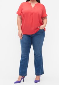 Blouse rouge à manches courtes avec un col en V et une coupe décontractée, associée à un jean bleu évasé et à des talons hauts violets. Texture de tissu lisse.